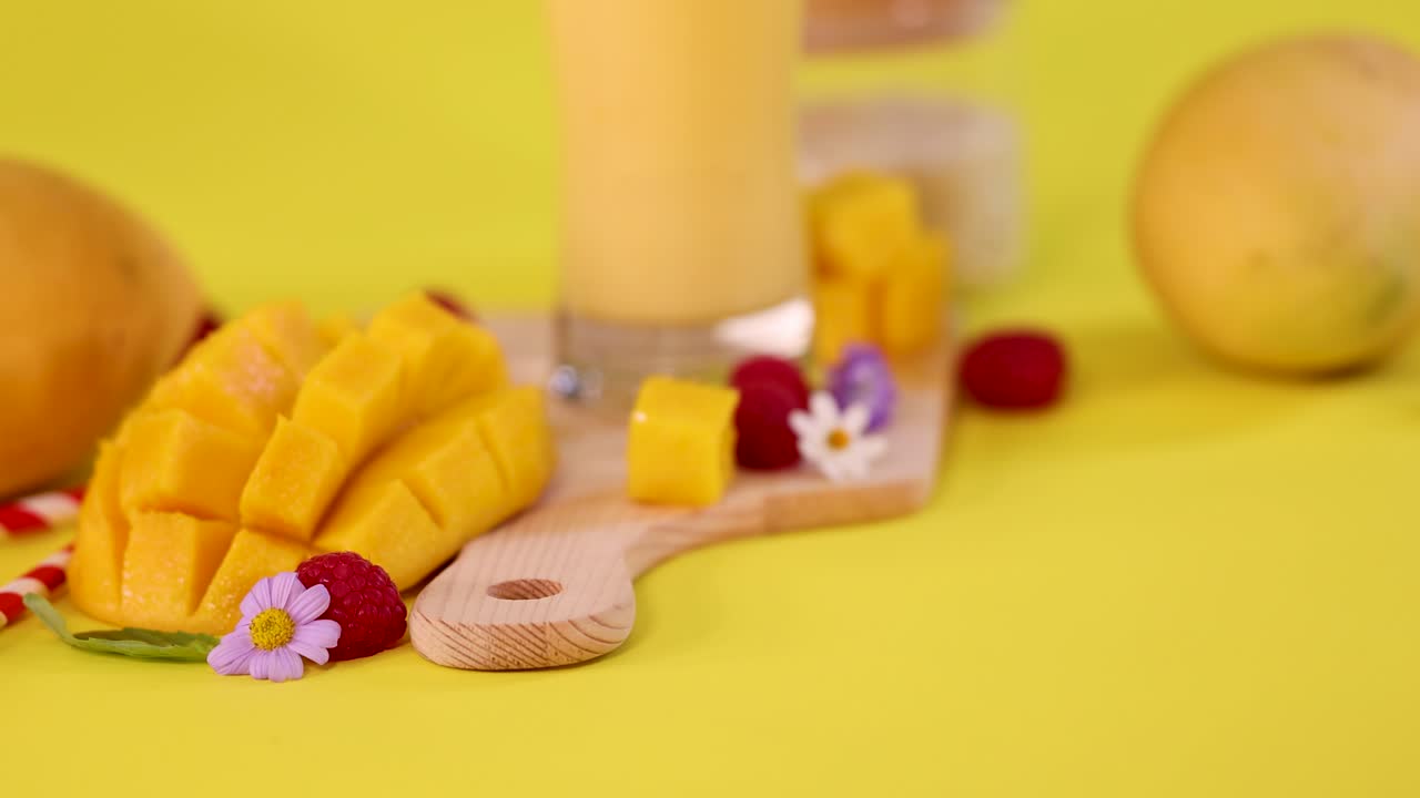 Spoon lifts whipped cream near mango cubes, smoothie, raspberries, and flowers on yellow background