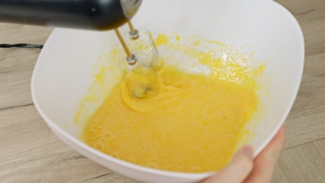hand mixer whips bright yellow egg yolks and sugar powder in a white bowl, blending ingredients smoothly for a cake or dessert base, fast spinning motion visible, real time overhead, making tiramisu