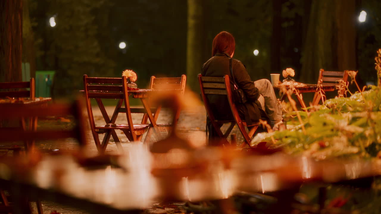 Rain gently falls in a peaceful park, creating a serene atmosphere. A lone person sits on a wooden chair, surrounded by wet foliage and dim lights in the evening