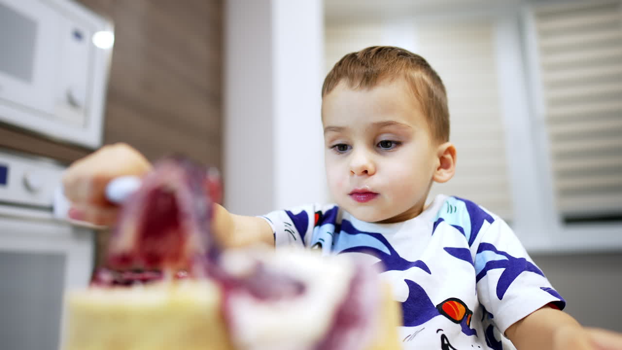 Beautiful toddler boy puts a fork into the cake. Kid takes away the jelly top and tastes the dessert. Low angle view.