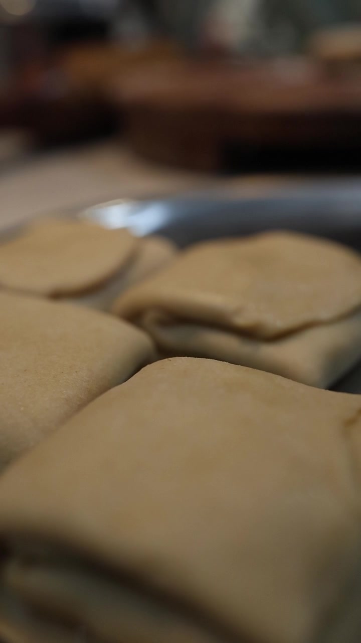 Close-up of hands kneading and folding soft chapati dough on a wooden surface in a home kitchen. The pale, smooth dough is worked with careful technique, creating layered folds. Warm, natural lighting