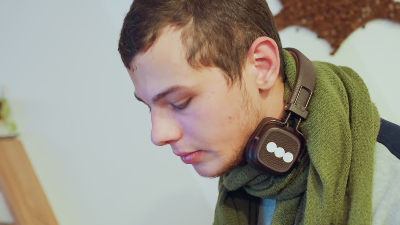 Joven blanco con auriculares llamando, probando niveles de audio y contestando el smartphone, bufanda verde, apartamento interior, retratos de primer plano, expresión concentrada, estilo invernal casual, cálido y suave