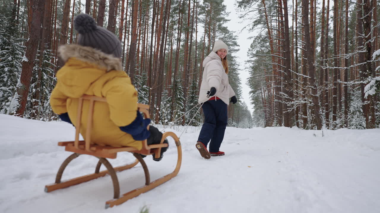 Glad ung kvinne går tur med sitt lille barn i vinterskogen, sønnen sitter i en trekjelke.
