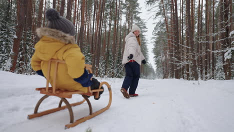 happy young woman is walking with her little child in winter forest son is sitting in wooden sleigh