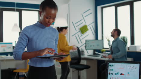 Portrait-of-african-american-woman-standing-in-busy-office-picking-up-tablet-with-business-erp-software-and-smiling