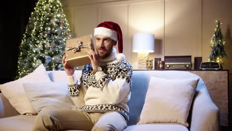 Alegre-Joven-Apuesto-De-Buen-Humor-Sonriendo-Con-Regalo-Envuelto-Sentado-En-Una-Casa-Decorada-Cerca-Del-árbol-De-Navidad-Con-Luces-Centelleantes-En-La-Víspera-De-Navidad