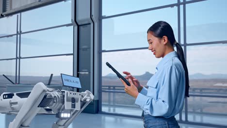 vista lateral de una trabajadora profesional asiática de pie con su tableta en una fábrica de alta tecnología, perro robot, comprobando su tableta con meditación