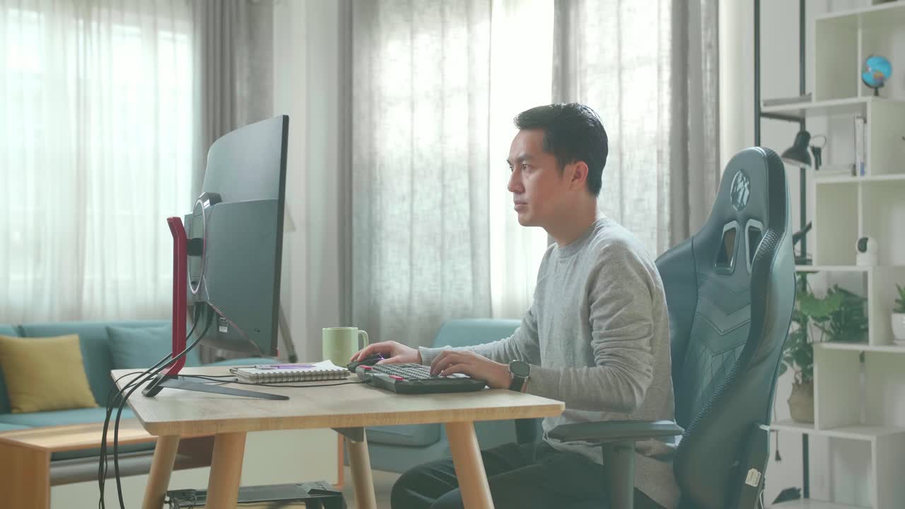 Asian Man In Long Sleeved T-Shirt And Black Pants Using Desktop Computer On A Table For Working At Home.