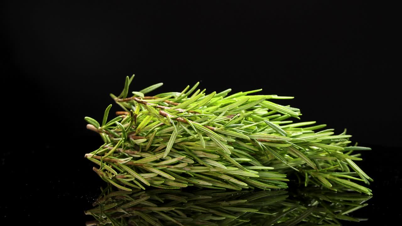 A bundle of rosemary rotates smoothly on a reflective black surface, highlighting its texture and vibrant green color