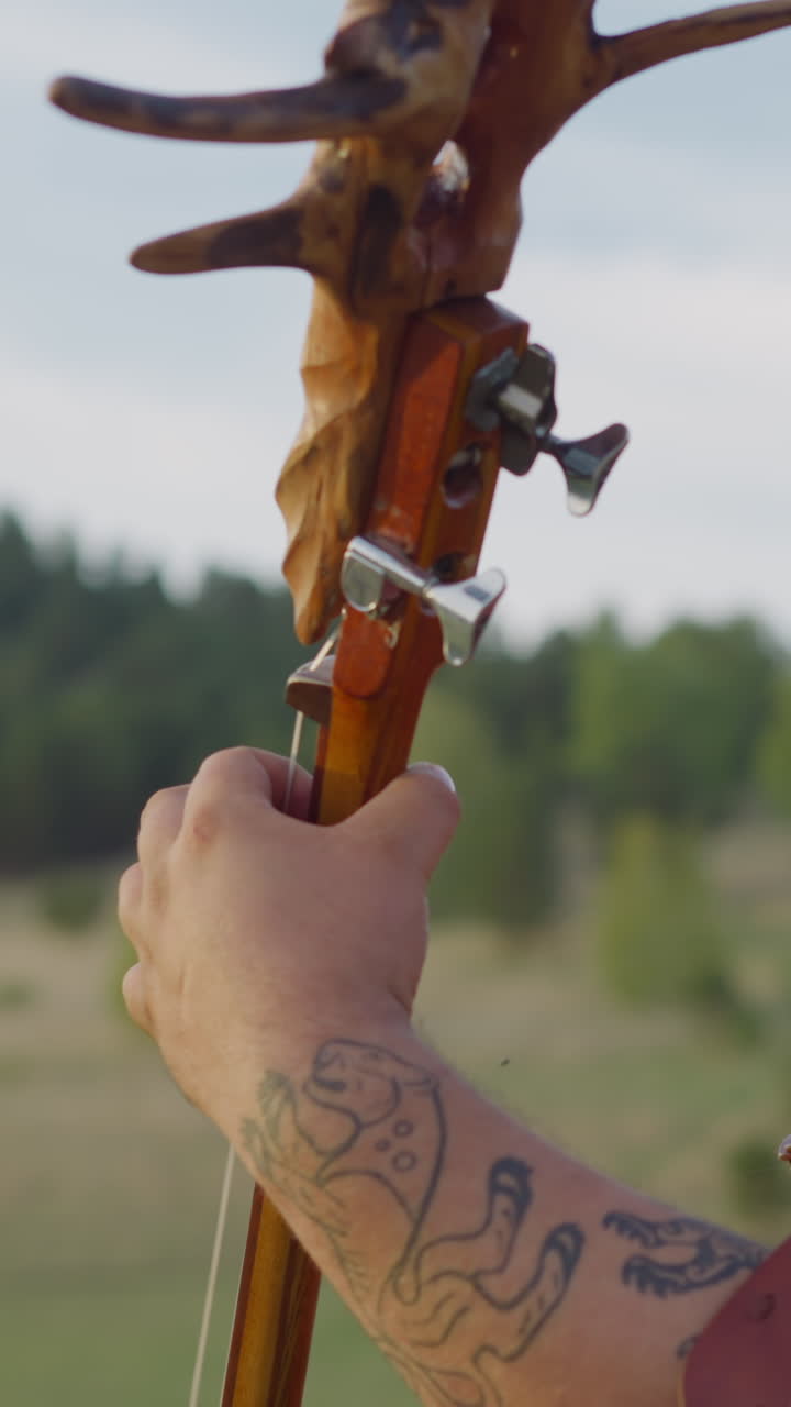 Obese bearded guy in leather Altai costume plays ikili decorated with roots in valley close side view. Participant of traditional culture fest