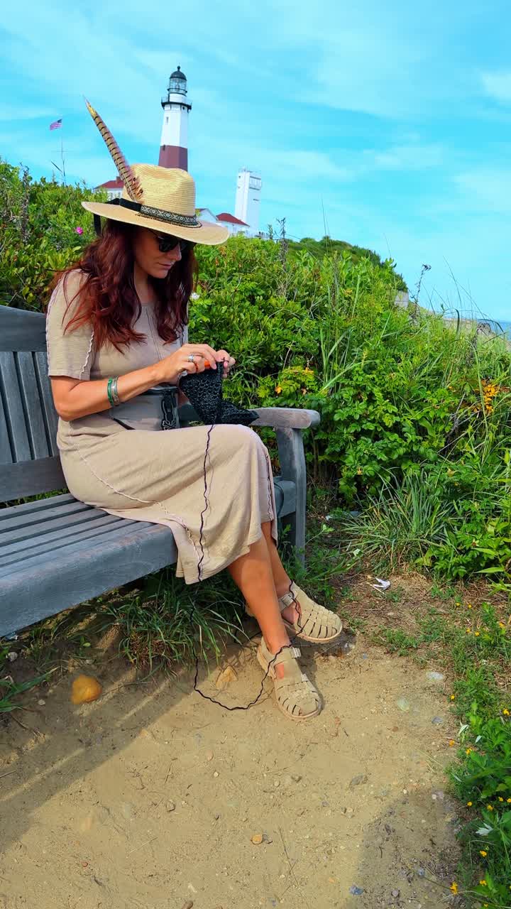 Donna con cappello di paglia che lavora a maglia su una panchina vicino al faro. Donna adulta in abito e cappello da sole che lavora a maglia filato nero mentre è seduta su una panchina di legno all'aperto