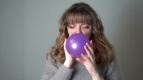 mujer joven que sopla un globo púrpura contra un fondo gris