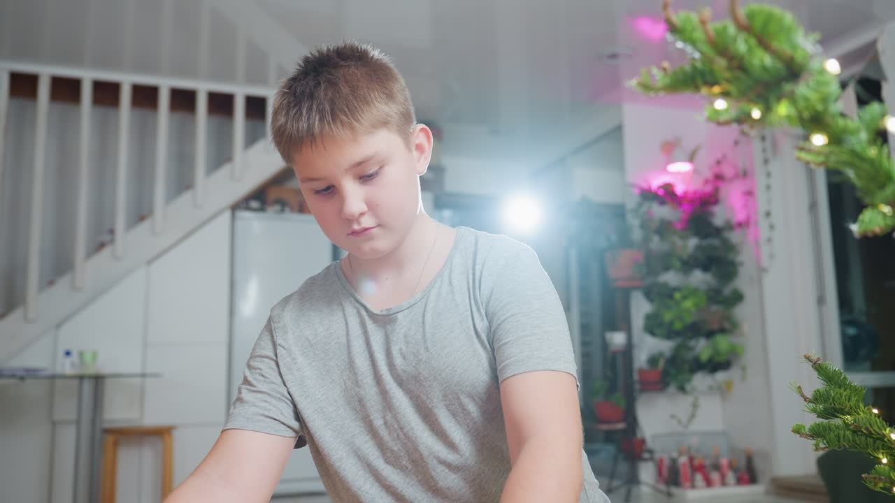 Boy in grey top carefully opening vintage box beside Christmas tree with glowing lights, cozy decorated indoor scene featuring colorful flowers and blurred figure of woman sitting behind