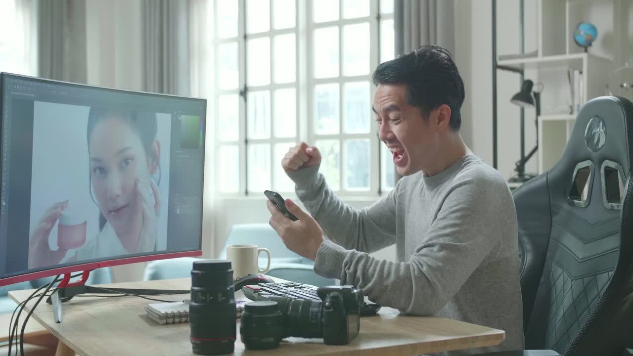 Asian Cameraman In Long Sleeved T-Shirt Looking At Mobile Phone And Being Happy Due To Success While Using Desktop Computer For Working At Home.
