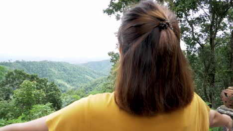 woman facing the forest from a high view deck her back on camera lifting arms with open palms forming a cross enjoying the refreshing nature