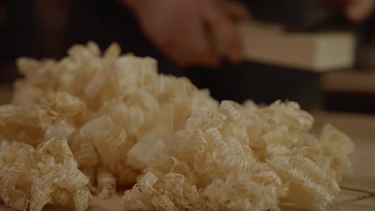Detailed close-up of fresh wood shavings on a workbench with a blurred background of a craftsman working, symbolizing woodworking, craftsmanship, and handmade furniture creation