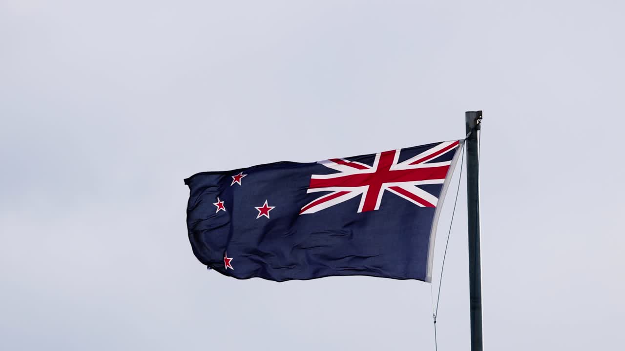 The New Zealand national flag flutters vigorously atop a flagpole against an overcast sky, captured in steady, medium shots with natural, diffused daylight