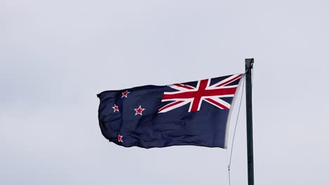 the new zealand national flag flutters vigorously atop a flagpole against an overcast sky, captured in steady, medium shots with natural, diffused daylight