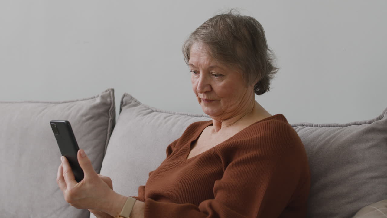 Senior Woman Using Smartphone While She Is Sitting On Sofa In Living Room At Home