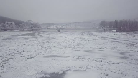 Drone-flying-towards-swing-bridge-above-a-snowy-river-with-big-ice-chunks-from-a-winter-storm