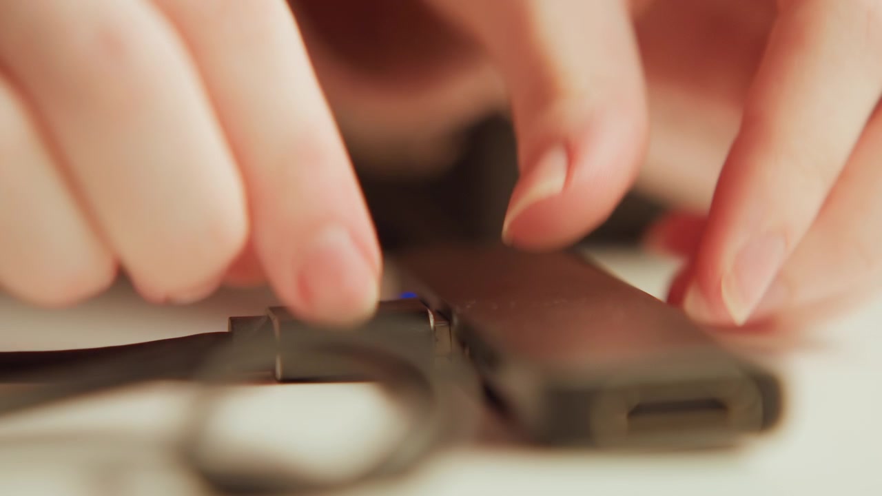 Extreme close-up of hands inserting USB cable into compact rectangular hub or external drive on light tabletop; fingers guide plug into port with subtle motion blur showing quick tactile movement