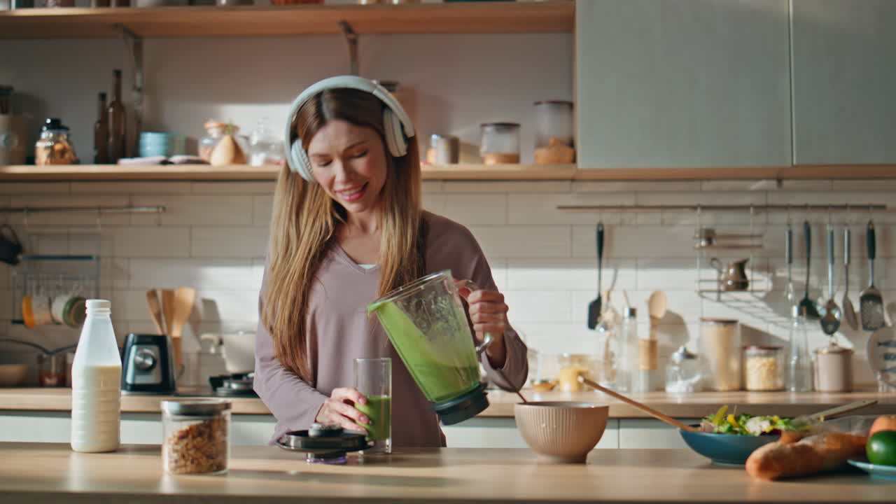 Dancing woman preparing smoothie listening music in headphones at cozy kitchen.