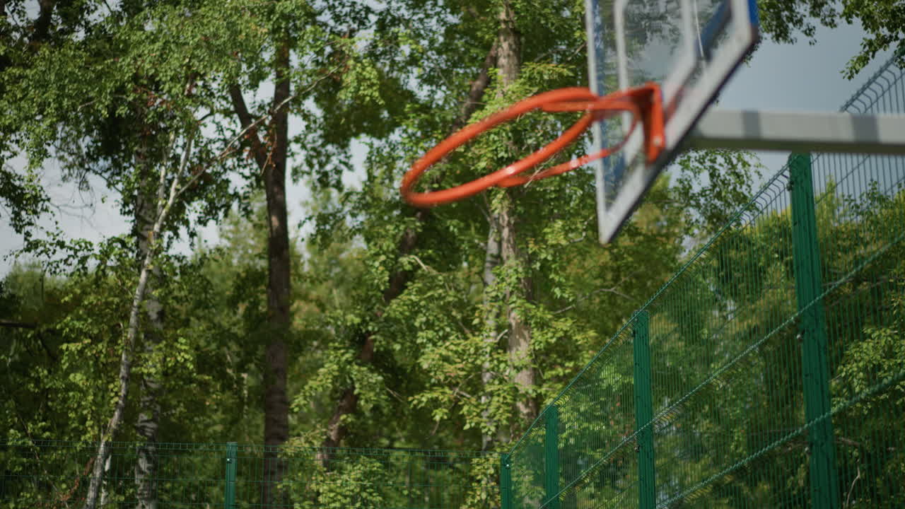 White Boy Shoots Soccer Ball Outdoors On Community Court, Dynamic Shot Framed By Hoop And Trees, Playful Crossover Of Soccer And Basketball Skills, Athletic Youth Practice Under Summer Canopy