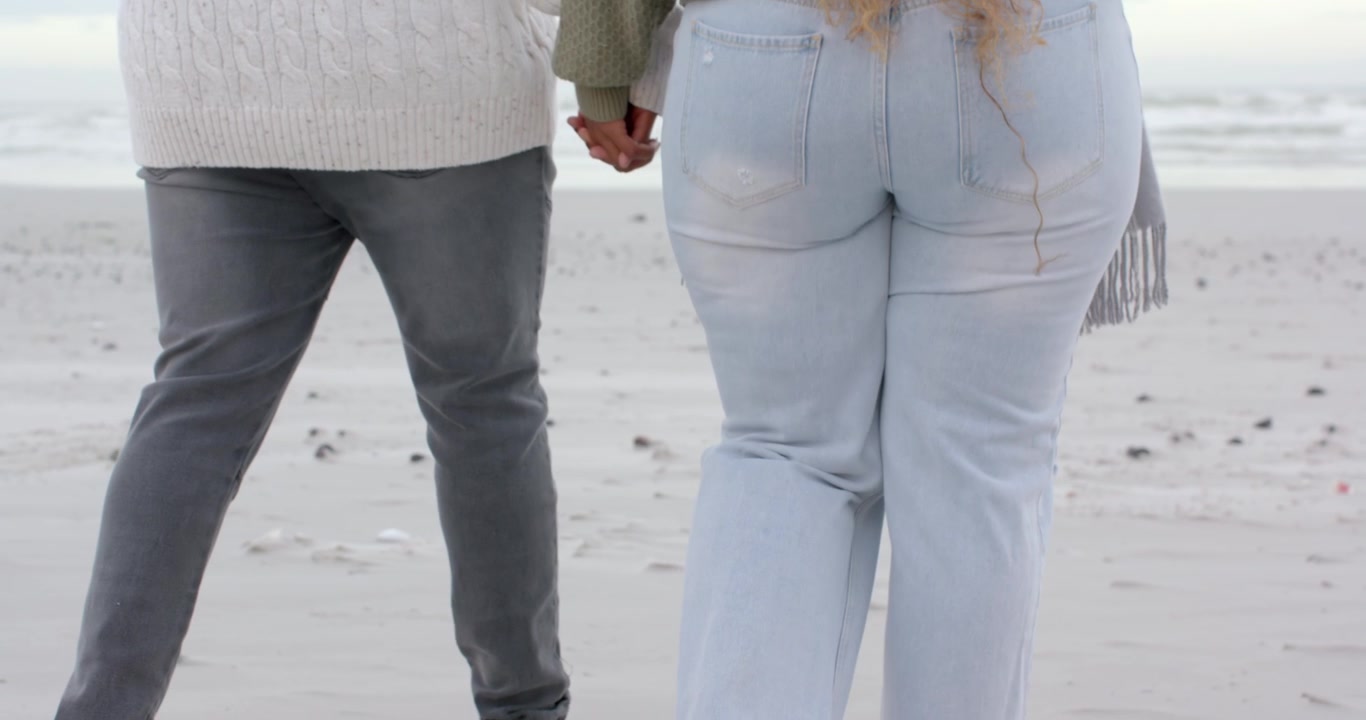 Couple walking onto pale sand, boots and sneakers leaving footprints while holding hands toward sea
