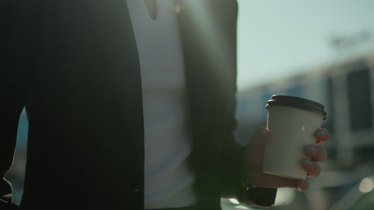 Close up of young woman in business attire walking outdoors holding tea cup, sunlight casting glow on her hand with blurred background of cityscape, bokeh lights, parked cars, and urban reflections