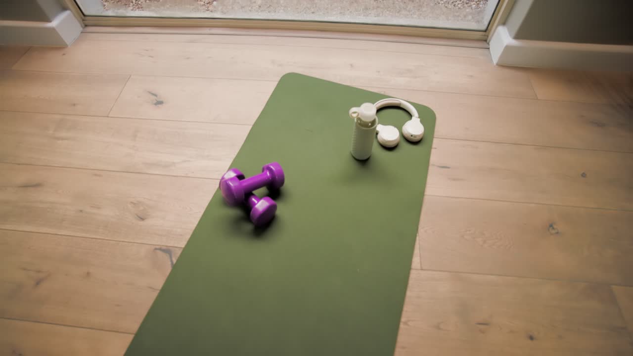La cámara se desliza hacia una esterilla de yoga verde, mostrando pesas moradas, una botella blanca y unos auriculares para hacer ejercicio.