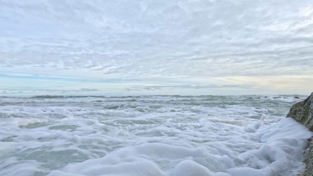 Dynamic ocean waves crash against rocky shores, creating foamy splashes under a cloudy sky