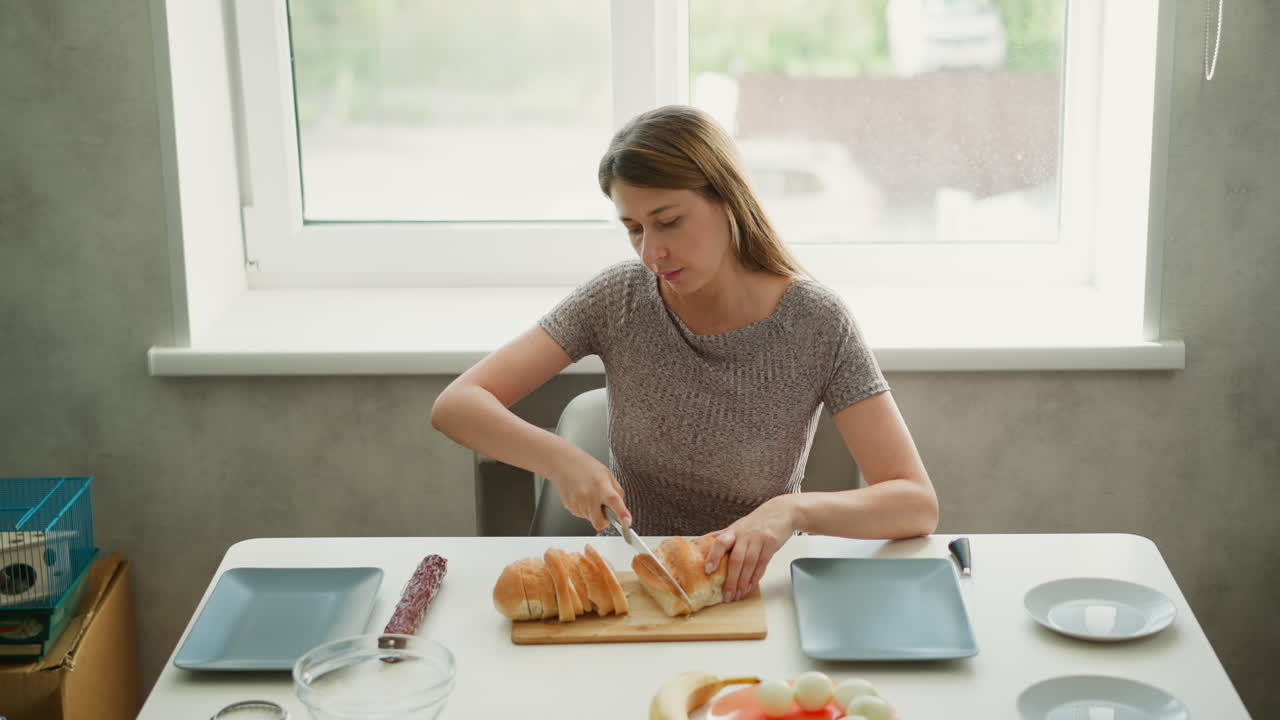 High angle shot of young baker cutting loaf of bread with knife on wooden board at kitchen table with eggs and white bowls under bright sunny morning light streaming through window behind