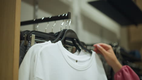 hand wearing pink cloth reaches for a white top from a clothing rack, ash trouser is seen behind in a brightly lit store, with a blurred background showing other clothes neatly arranged on shelves