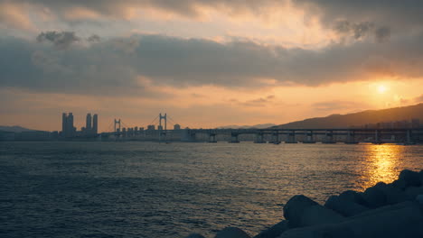 gwangan bridge with dramatic sky during golden sunset in busan, sun setting over hwangnyeongsan mountain - wide angle