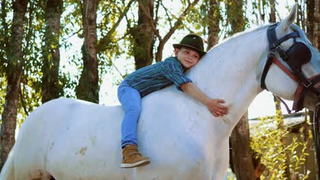 fotografía auténtica de un niño lindo con un sombrero está acostado en un caballo blanco y acariciando su