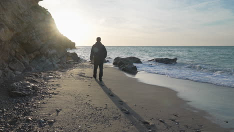 man in black jacket walks on beach, leaving footsteps behind as well long shadow, during sunny morning, sea waves wash onshore