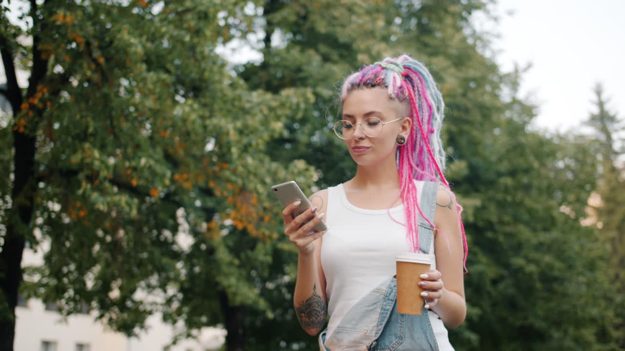 Stylish young woman with colorful dreadlocks, wearing denim overalls, walking in a park and using her phone.
