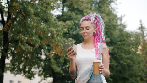 stylish young woman with colorful dreadlocks, wearing denim overalls, walking in a park and using her phone.