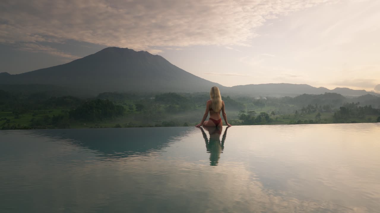 Fitte Frau im Bikini am Poolrand blickt auf einen magischen Sonnenaufgang mit dem Vulkan Agung.