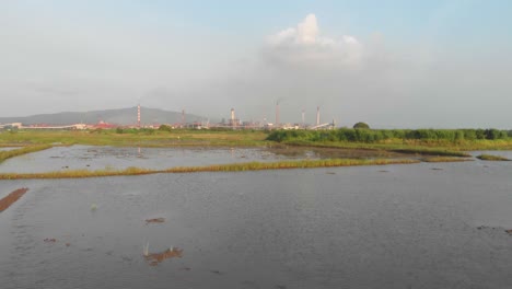 low slider drone shot over waterlogged indian rice paddy farming fields large factory chimneys background