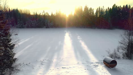 Dron-Aéreo-En-Movimiento-Hacia-Atrás-De-Un-Amanecer-Brillante-Desde-Detrás-De-árboles-Coníferos-Sobre-Un-Lago-Congelado-Junto-A-Una-Sauna-De-Barril