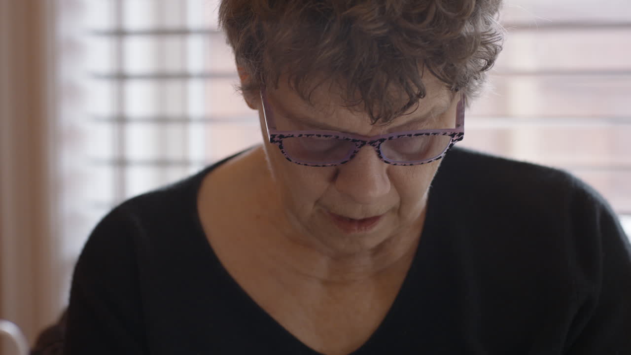 An elderly woman's face focused on gently self-massaging the spasticity in her hemiplegic right hand. Seated by round glass table with legs in checkered pants, Camera tilts down from face to hands.