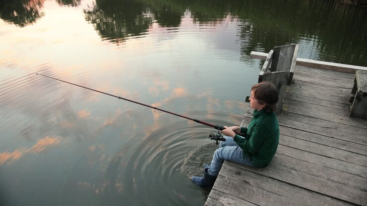 High angle view of a little boy fishing while he is sitting on the lake pier at sunset