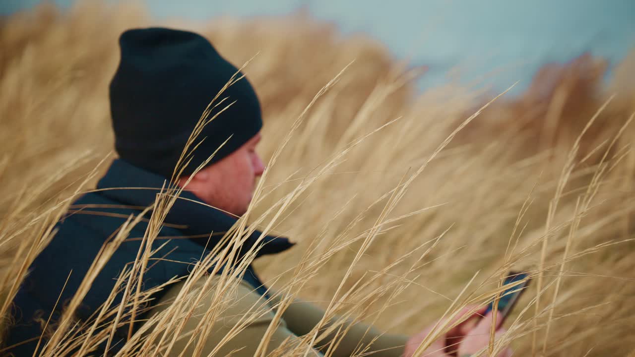 Een nadenkend figuur, zittend te midden van lang, goudkleurig gras, toont een sereen moment van introspectie, terwijl zacht zonlicht door de grassprieten filtert en een rustige en reflecterende sfeer creëert