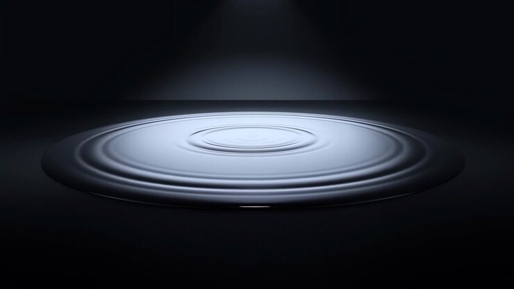 Circular waves expand outward from small central disturbance across silvery blue liquid, with shifting highlights and dark edges framing smooth reflective surface beneath overhead spotlight.