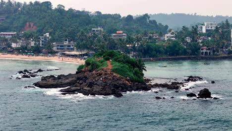 tropical sri lanka slightly elevated view of a rocky islet covered in lush green vegetation, near a sandy beach and coastal town under an overcast, humid sky, captured with a standard lens