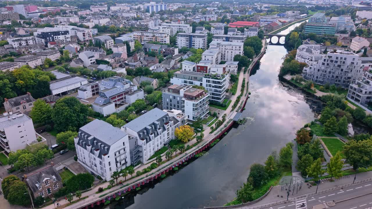 Drone fly over the Vilaine River flowing between residential buildings and green areas with railway bridge in the background, Rennes, Ille-et-Vilaine, France