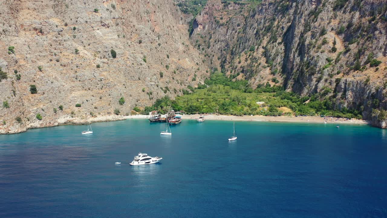 aerial drone panning across the white sand beach of Butterfly Valley in Fethiye Turkey with boats anchored in the bay and blue ocean on a sunny summer day