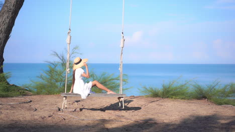 amazing asian woman gently sitting on the big wooden bench swing wearing summer sundress and hat on blue sea background in thailand, slow motion copy space