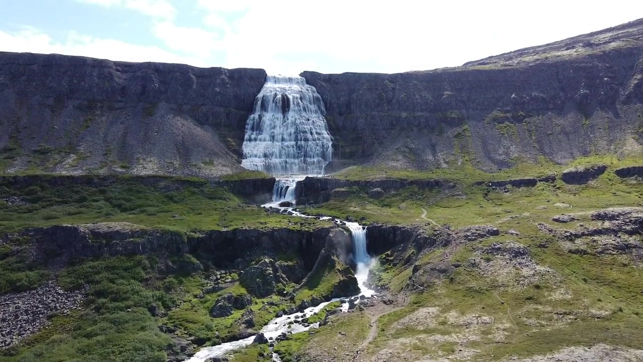 Il filmato aereo mostra una strada serpeggiante che costeggia il fiume Skjálfandafljót e le cascate superiori mentre si gettano nella gola di Goðafoss. La luce del giorno nitida rivela un terreno lussureggiante e una cascata dinamica.